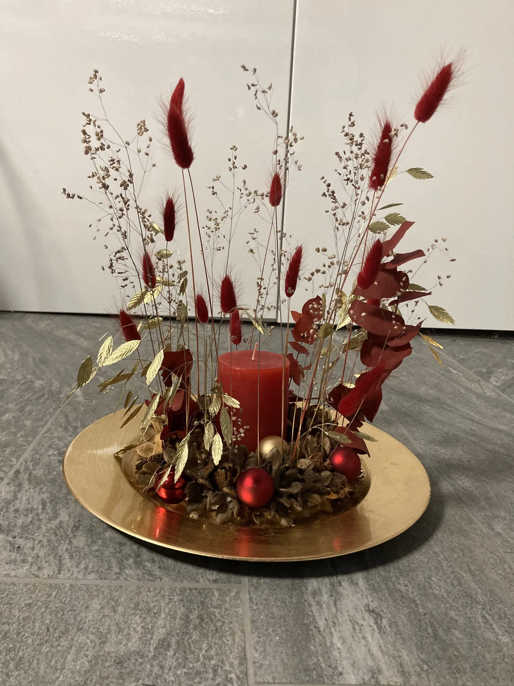 Adventsgesteck auf goldener Schale mit roter Kerze und roten Trockengräsern