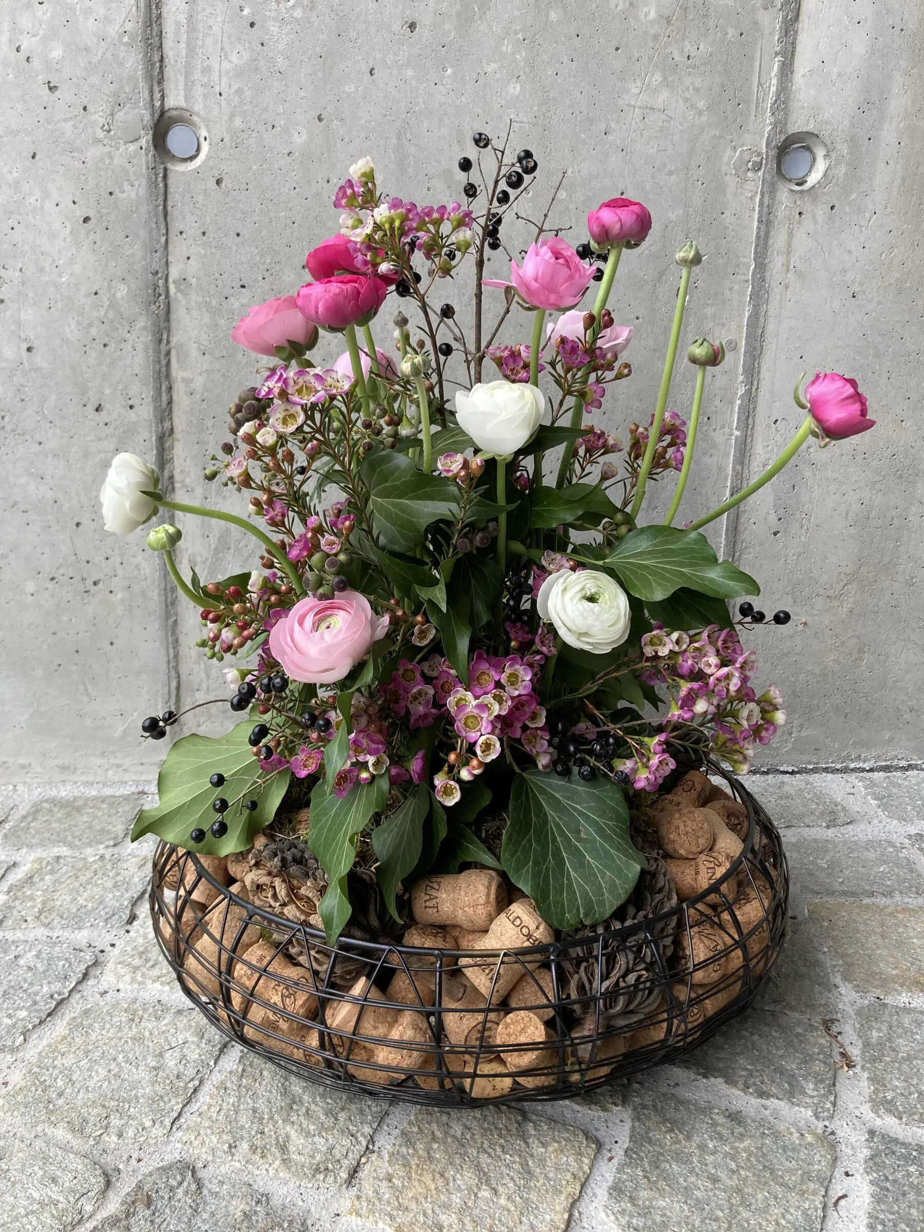 Drahtkorb mit Korken, pinken Ranunkeln und Wachsblumen