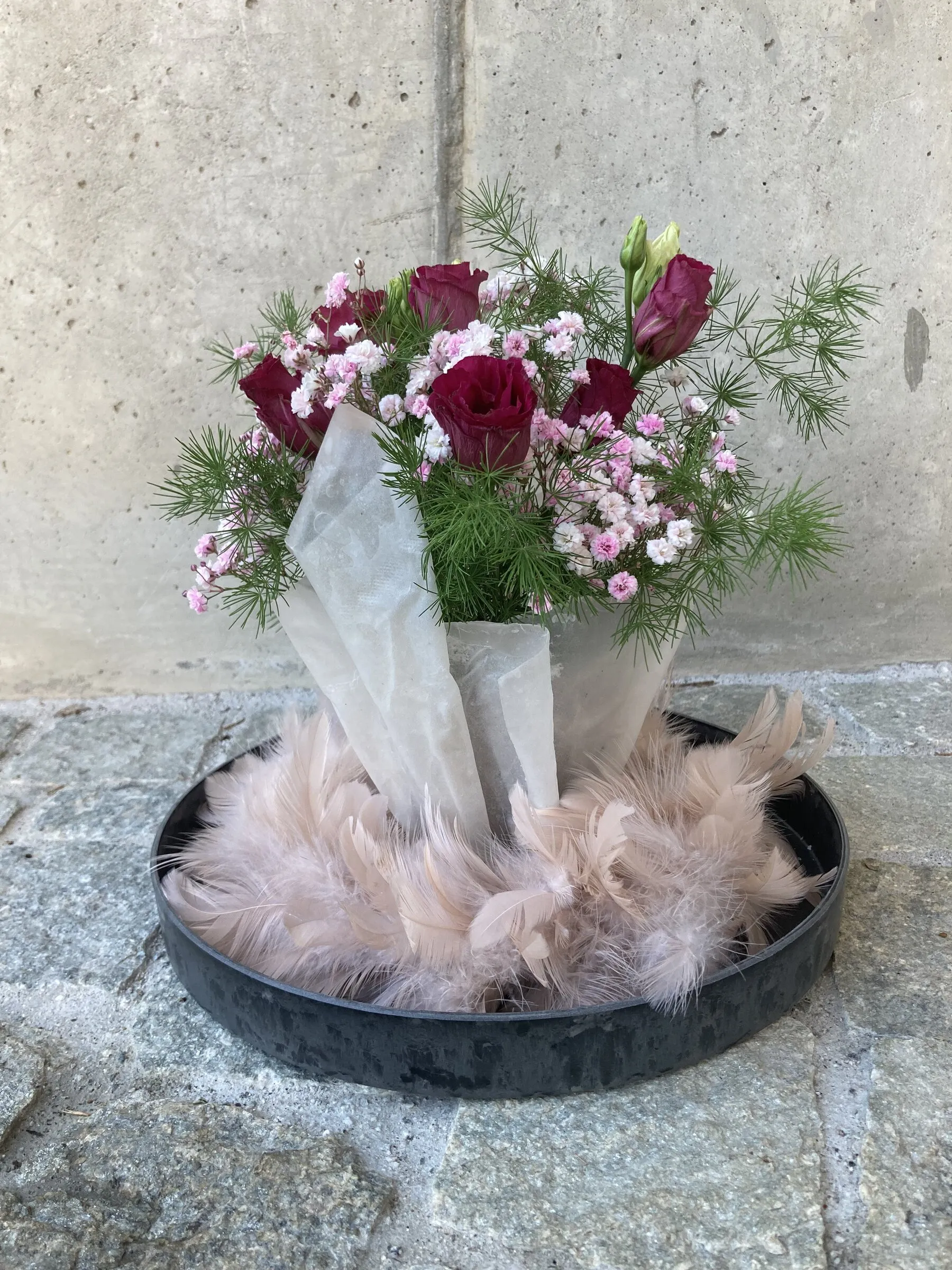 Dunkelrote Rosen in Papiertüten auf einer Schale mit rosa Federn