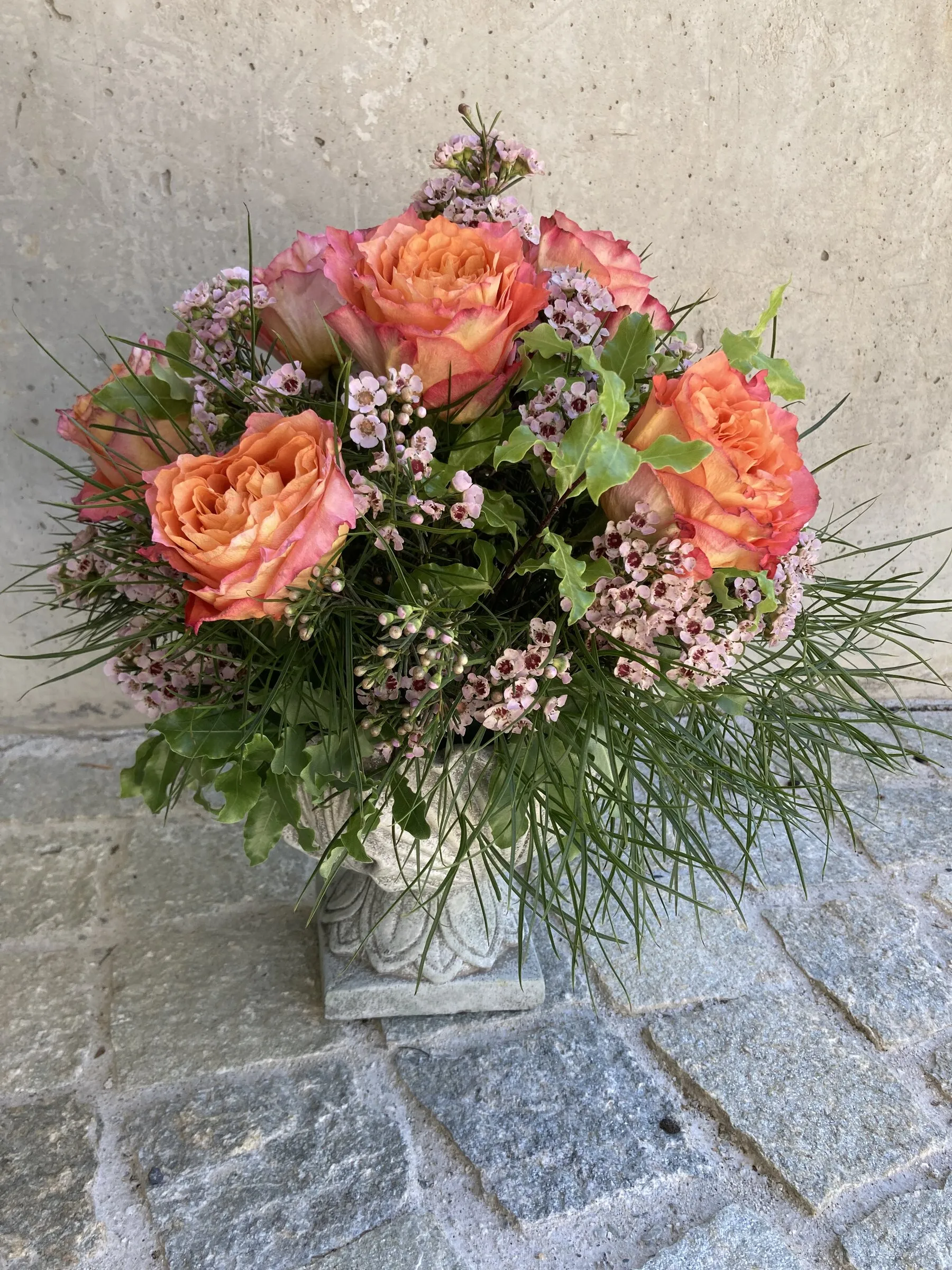 Steinvase mit orangen Rosen, rosa Wachsblumen und Gräsern