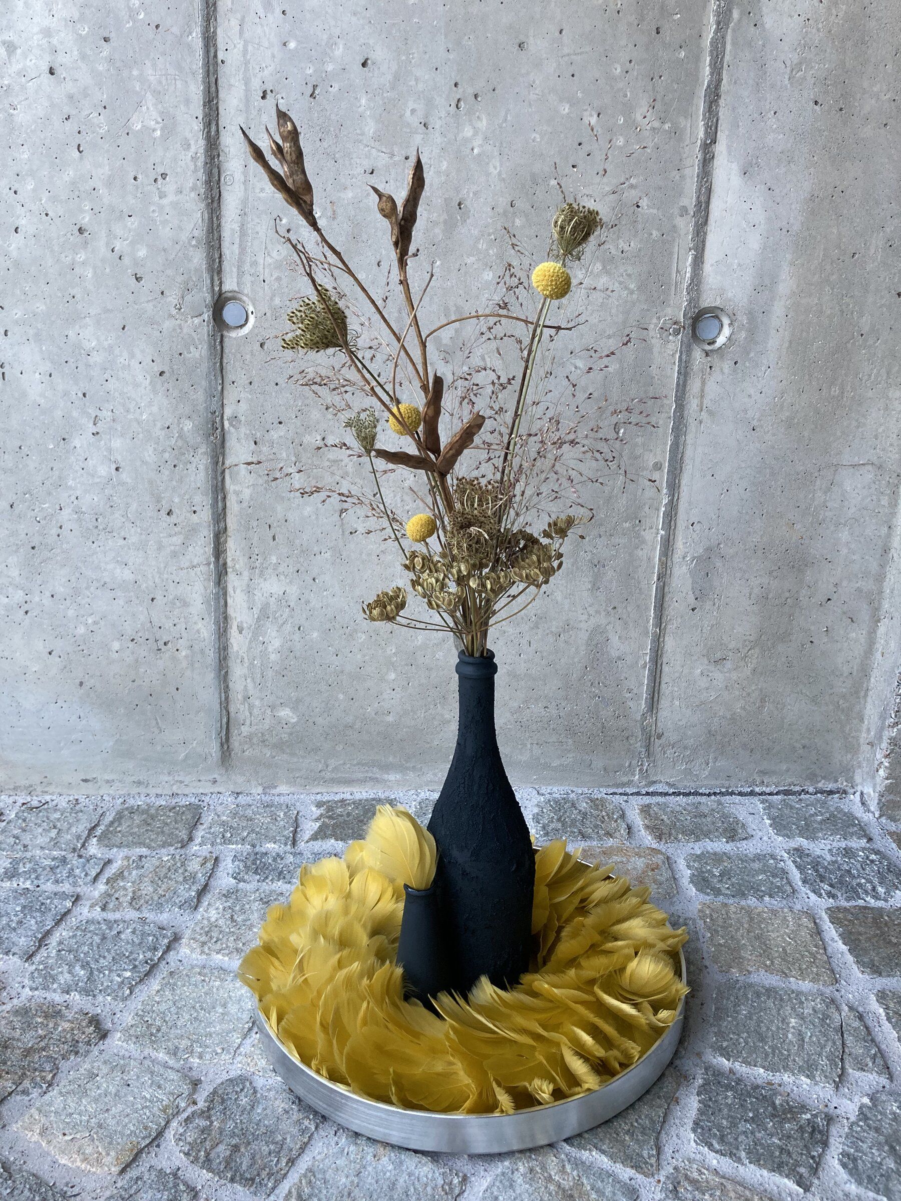 Schwarze Vase mit Trockengräsern auf einem gelben Federkranz